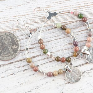 May include: A pair of silver hoop earrings with a hammered silver disc charm. The hoops are adorned with multi-colored gemstone beads.