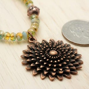 Pu&ograve; includere: Una collana con pendente color rame con un grande e dettagliato motivo floreale. La collana ha un filo di perline con perline gialle, verdi e blu. Una moneta &egrave; accanto alla collana come riferimento delle dimensioni.