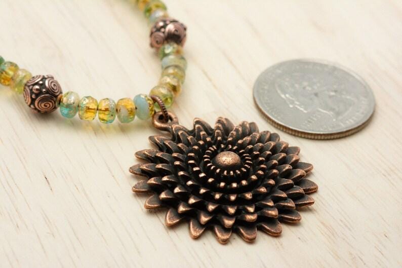 Pu&ograve; includere: Una collana con pendente color rame con un grande e dettagliato motivo floreale. La collana ha un filo di perline con perline gialle, verdi e blu. Una moneta &egrave; accanto alla collana come riferimento delle dimensioni.