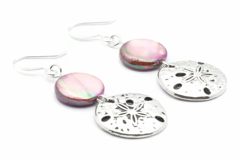 Puede incluir: Un par de pendientes de plata con una concha rosa iridiscente y un dije de d&oacute;lar de arena plateado.