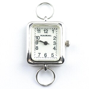 Puede incluir: Un reloj de bolsillo plateado con una esfera rectangular y una esfera blanca. El reloj presenta n&uacute;meros y manecillas negros, con el nombre de la marca "NARMI" impreso en el centro. Se adjuntan dos anillos plateados para colgar.