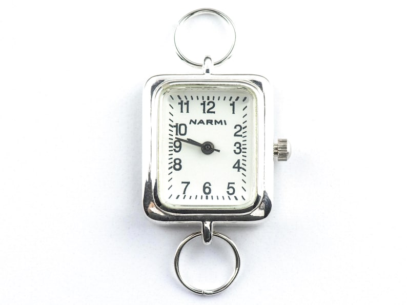 Puede incluir: Un reloj de bolsillo plateado con una esfera rectangular y una esfera blanca. El reloj presenta n&uacute;meros y manecillas negros, con el nombre de la marca "NARMI" impreso en el centro. Se adjuntan dos anillos plateados para colgar.