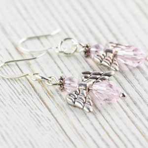 May include: A pair of silver dangle earrings with pink crystal angel charms. The earrings have a hook closure.