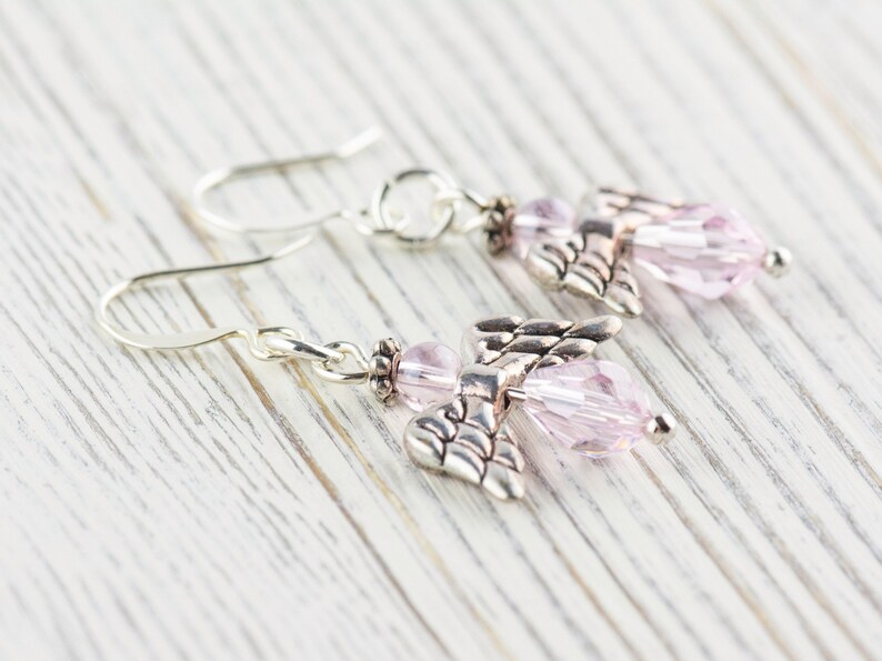 May include: A pair of silver dangle earrings with pink crystal angel charms. The earrings have a hook closure.