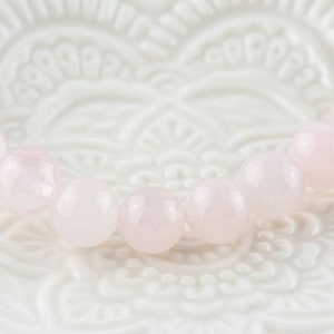 Puede incluir: Un brazalete de cuarzo rosa con cuentas redondas. El brazalete est&aacute; sobre un fondo blanco con un dise&ntilde;o floral.