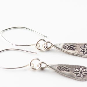 Puede incluir: Un par de pendientes de plata con un dije en forma de l&aacute;grima que presenta un dise&ntilde;o de flor estilizada y flecha.