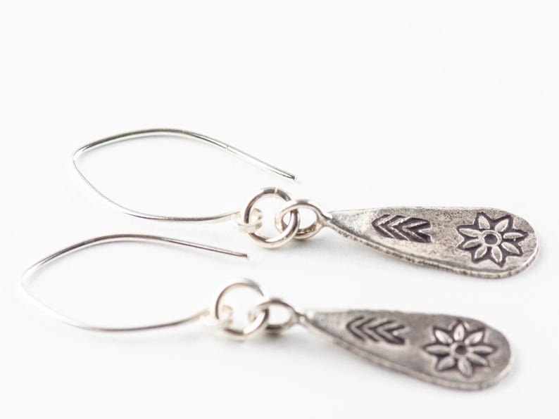 Puede incluir: Un par de pendientes de plata con un dije en forma de l&aacute;grima que presenta un dise&ntilde;o de flor estilizada y flecha.