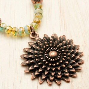 Pu&ograve; includere: Una collana con pendente a forma di girasole color rame con una catena di perline gialle, verdi e blu. Il pendente presenta petali dettagliati e un motivo a spirale centrale. La collana &egrave; un gioiello.
