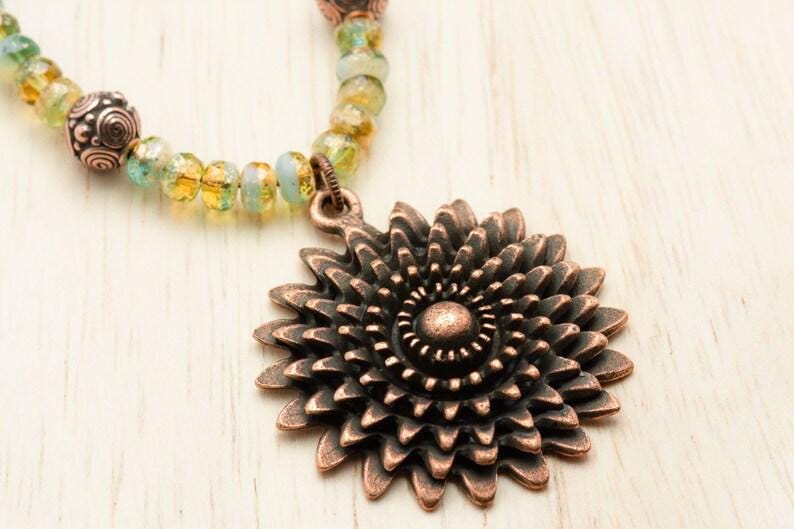 Pu&ograve; includere: Una collana con pendente a forma di girasole color rame con una catena di perline gialle, verdi e blu. Il pendente presenta petali dettagliati e un motivo a spirale centrale. La collana &egrave; un gioiello.