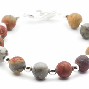 May include: A beaded bracelet featuring round, multicolored jasper stones in shades of red, brown, gray, and tan. The beads are separated by small, silver-colored metal beads and connected by a silver-colored clasp.