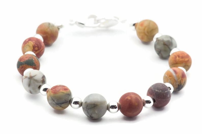 May include: A beaded bracelet featuring round, multicolored jasper stones in shades of red, brown, gray, and tan. The beads are separated by small, silver-colored metal beads and connected by a silver-colored clasp.