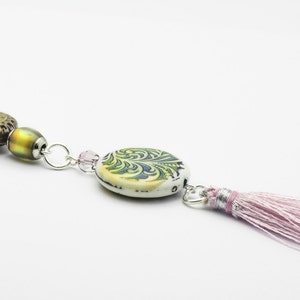 May include: A delicate necklace with a pink tassel, featuring a green and white patterned ceramic bead, a brown and green patterned ceramic bead, and a clear iridescent glass bead.