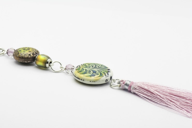 May include: A delicate necklace with a pink tassel, featuring a green and white patterned ceramic bead, a brown and green patterned ceramic bead, and a clear iridescent glass bead.