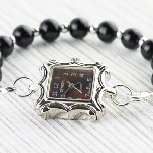 May include: A silver watch with a square face and black numbers. The watch is attached to a black beaded bracelet with a silver clasp.