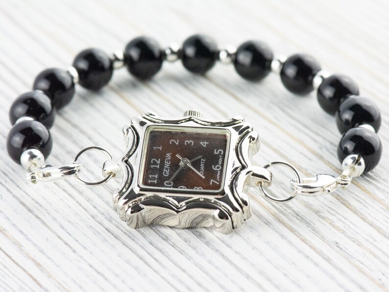 May include: A silver watch with a square face and black numbers. The watch is attached to a black beaded bracelet with a silver clasp.