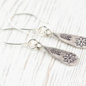 Puede incluir: Un par de pendientes de plata con forma de l&aacute;grima. Cada pendiente tiene un peque&ntilde;o dise&ntilde;o de flor y una serie de tres flechas.