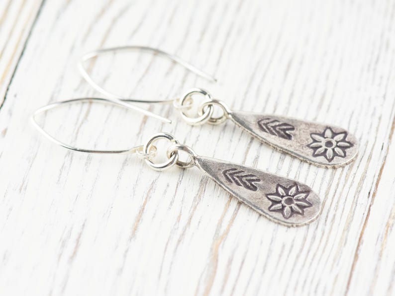 Puede incluir: Un par de pendientes de plata con forma de l&aacute;grima. Cada pendiente tiene un peque&ntilde;o dise&ntilde;o de flor y una serie de tres flechas.