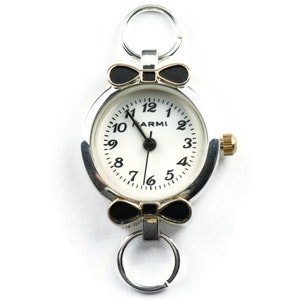 May include: A silver-toned watch with a white face and black numbers. The watch has a black bow on either side of the face and a silver ring on the top. The watch face says "ARMI".