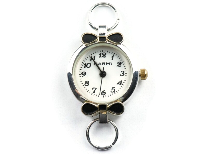 May include: A silver-toned watch with a white face and black numbers. The watch has a black bow on either side of the face and a silver ring on the top. The watch face says "ARMI".