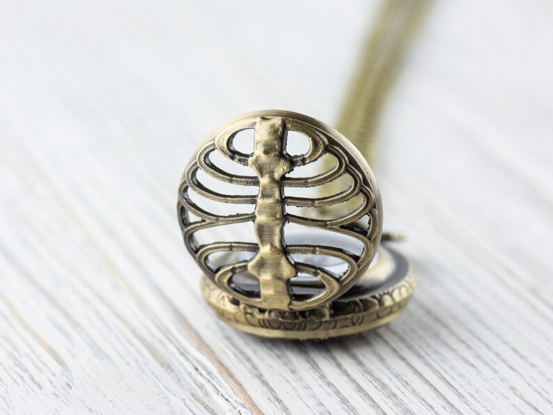 May include: A gold-toned necklace with a round pendant featuring a detailed ribcage design. The pendant is open and has a small glass window inside.