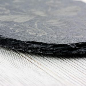 May include: A round, dark gray slate coaster with a floral design etched into the surface. The coaster has a rough, natural edge and sits on a white wooden surface. The coaster is a decorative item for the home.