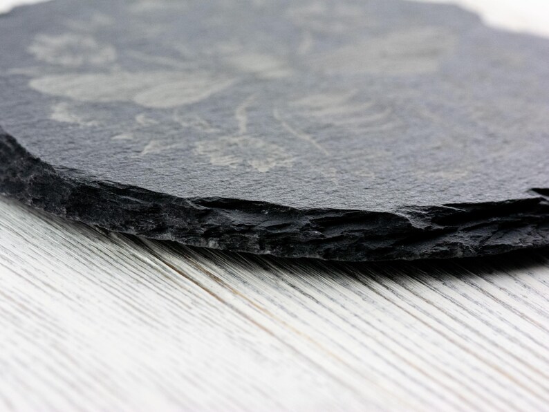 May include: A round, dark gray slate coaster with a floral design etched into the surface. The coaster has a rough, natural edge and sits on a white wooden surface. The coaster is a decorative item for the home.