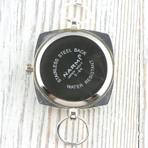May include: A silver watch with a square face and a black dial. The watch back is inscribed with "STAINLESS STEEL BACK," "NARMI JAPAN MOVT S-64," and "WATER RESISTANT." It features a small crown and a ring for attachment.