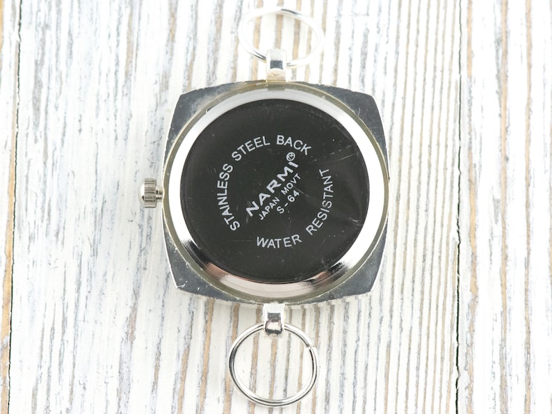 May include: A silver watch with a square face and a black dial. The watch back is inscribed with "STAINLESS STEEL BACK," "NARMI JAPAN MOVT S-64," and "WATER RESISTANT." It features a small crown and a ring for attachment.