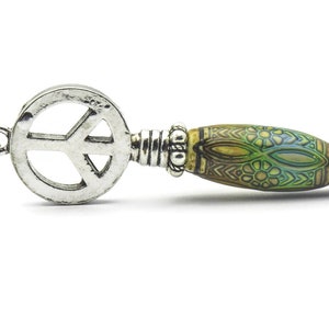 Puede incluir: Un colgante de plata con un s&iacute;mbolo de paz y una cuenta de color verde y marr&oacute;n con un patr&oacute;n, unido a una cadena de plata.
