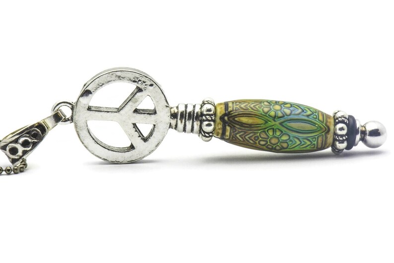 Puede incluir: Un colgante de plata con un s&iacute;mbolo de paz y una cuenta de color verde y marr&oacute;n con un patr&oacute;n, unido a una cadena de plata.