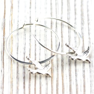 Peut inclure: Boucles d'oreilles cr&eacute;oles argent&eacute;es avec un charm en forme d'oiseau. Les boucles d'oreilles circulaires ont un petit pendentif oiseau attach&eacute;. Les oiseaux sont d&eacute;taill&eacute;s avec des ailes grav&eacute;es. Pr&eacute;sent&eacute;es sur une surface en bois blanc.