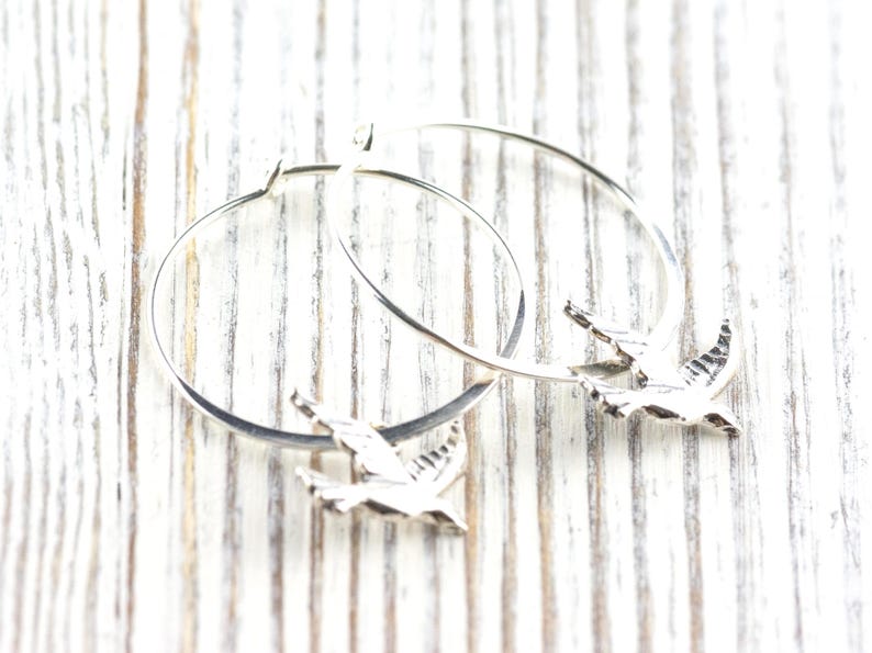 Peut inclure: Boucles d'oreilles cr&eacute;oles argent&eacute;es avec un charm en forme d'oiseau. Les boucles d'oreilles circulaires ont un petit pendentif oiseau attach&eacute;. Les oiseaux sont d&eacute;taill&eacute;s avec des ailes grav&eacute;es. Pr&eacute;sent&eacute;es sur une surface en bois blanc.