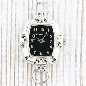 Pu&ograve; includere: Orologio da tasca argentato con quadrante nero e numeri bianchi. L'orologio ha una forma rettangolare con decorazioni e due anelli per l'attacco. Il marchio "NARMI" &egrave; visibile sul quadrante.