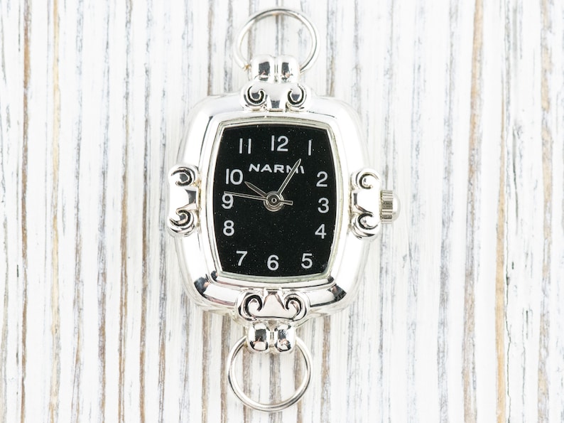 Pu&ograve; includere: Orologio da tasca argentato con quadrante nero e numeri bianchi. L'orologio ha una forma rettangolare con decorazioni e due anelli per l'attacco. Il marchio "NARMI" &egrave; visibile sul quadrante.