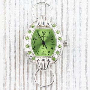 Op de afbeelding: Een zilveren horloge met een groene wijzerplaat en groene kristallen accenten. De wijzerplaat van het horloge zegt "Geneva Quartz" en heeft Romeinse cijfers voor de uren.