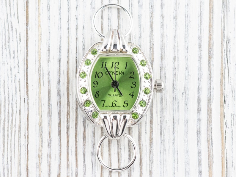 Op de afbeelding: Een zilveren horloge met een groene wijzerplaat en groene kristallen accenten. De wijzerplaat van het horloge zegt "Geneva Quartz" en heeft Romeinse cijfers voor de uren.