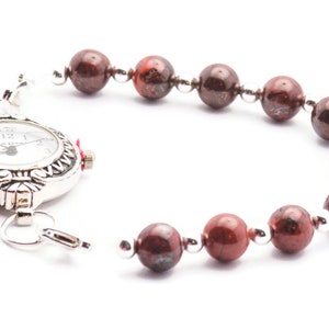May include: A silver pocket watch with a red stone accent attached to a bracelet made of red and brown beads.