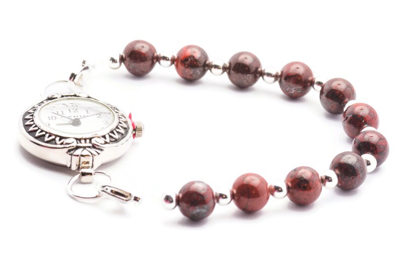May include: A silver pocket watch with a red stone accent attached to a bracelet made of red and brown beads.