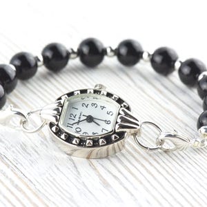 May include: A silver watch with a white face and black numerals is attached to a bracelet of large, glossy black beads. The watch has a decorative bezel with small, raised details. The bracelet is secured with silver clasps.