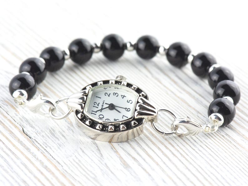May include: A silver watch with a white face and black numerals is attached to a bracelet of large, glossy black beads. The watch has a decorative bezel with small, raised details. The bracelet is secured with silver clasps.