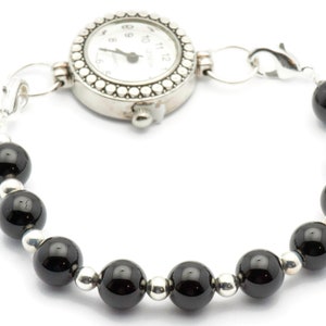 Puede incluir: Un brazalete de reloj plateado con una esfera redonda blanca y n&uacute;meros negros. La pulsera est&aacute; adornada con cuentas negras brillantes alternas y cuentas plateadas m&aacute;s peque&ntilde;as, conectadas por eslabones plateados.