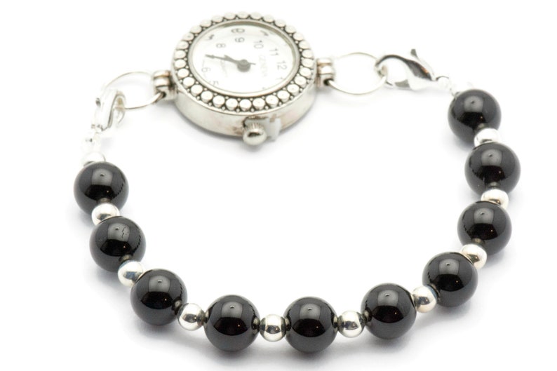 Puede incluir: Un brazalete de reloj plateado con una esfera redonda blanca y n&uacute;meros negros. La pulsera est&aacute; adornada con cuentas negras brillantes alternas y cuentas plateadas m&aacute;s peque&ntilde;as, conectadas por eslabones plateados.