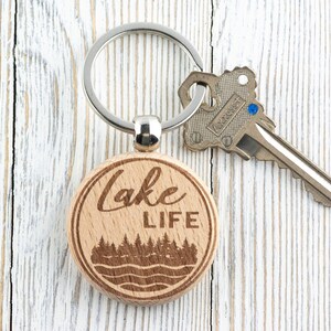 K&ouml;nnte beinhalten: Ein Holzschl&uuml;sselanh&auml;nger mit einem silbernen Ring und einem Schl&uuml;ssel. Der Schl&uuml;sselanh&auml;nger tr&auml;gt die Aufschrift "Lake LIFE" sowie ein Wald- und Wassermotiv. Der Schl&uuml;sselanh&auml;nger ist aus hellem Holz gefertigt.