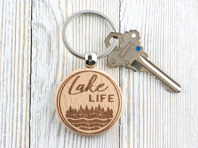 K&ouml;nnte beinhalten: Ein Holzschl&uuml;sselanh&auml;nger mit einem silbernen Ring und einem Schl&uuml;ssel. Der Schl&uuml;sselanh&auml;nger tr&auml;gt die Aufschrift "Lake LIFE" sowie ein Wald- und Wassermotiv. Der Schl&uuml;sselanh&auml;nger ist aus hellem Holz gefertigt.