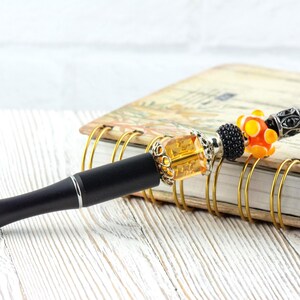 May include: A decorative pen with a black barrel and silver accents. The pen features a beaded design with orange, yellow, and black beads. The pen rests on a spiral-bound notebook with a floral design.