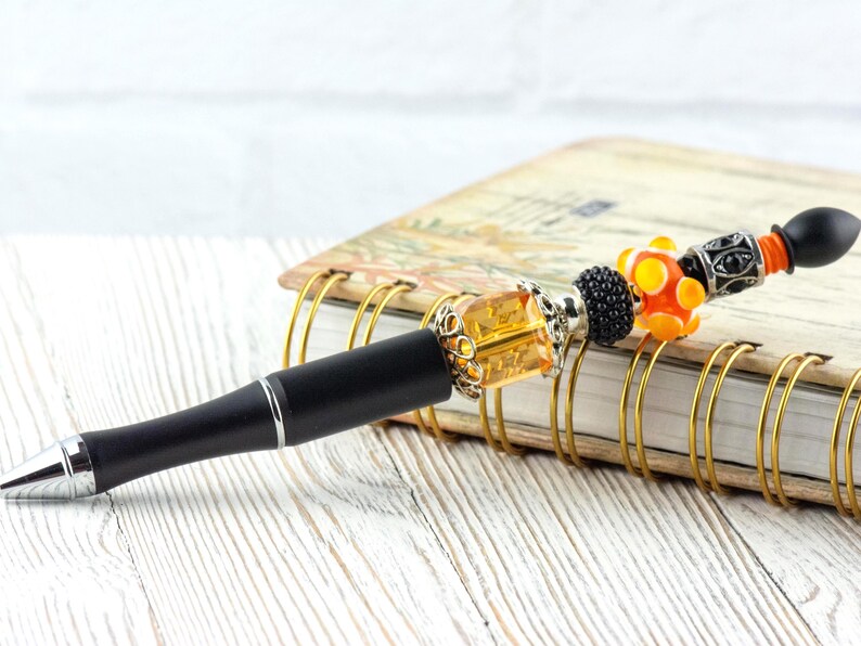 May include: A decorative pen with a black barrel and silver accents. The pen features a beaded design with orange, yellow, and black beads. The pen rests on a spiral-bound notebook with a floral design.
