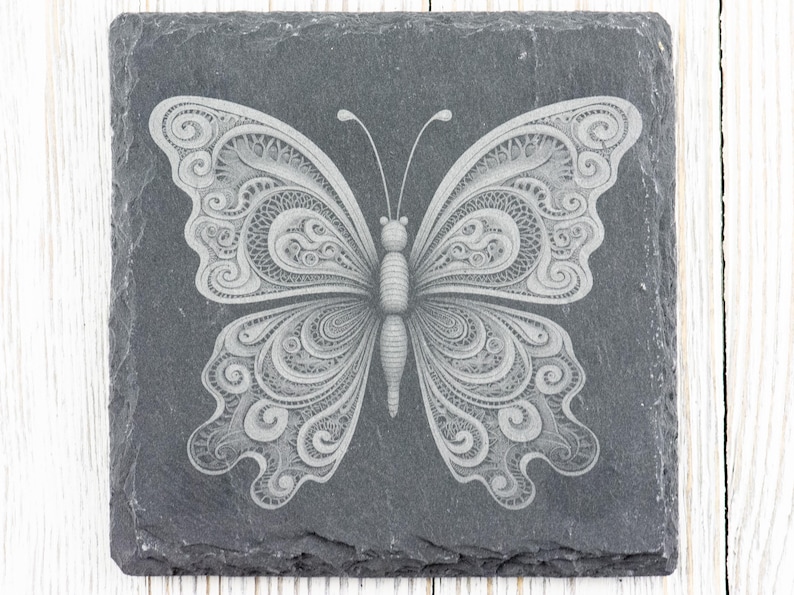 May include: A black slate coaster featuring a white, intricately engraved butterfly design.