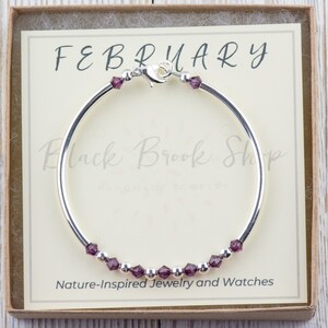 May include: A silver bangle bracelet with a purple crystal charm and the text "FEBRUARY" on a white card in a box. The card also says "Black Brook Shop Handmade in the UK" and "Nature-Inspired Jewelry and Watches".