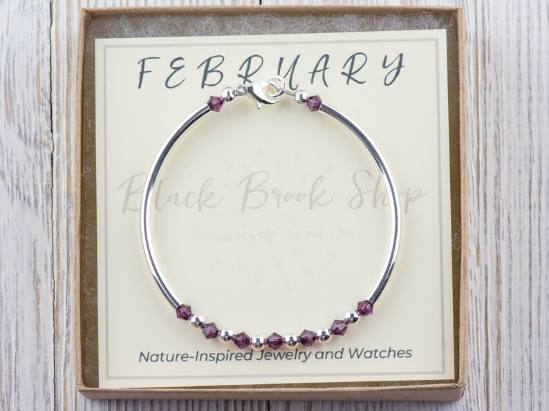 May include: A silver bangle bracelet with a purple crystal charm and the text "FEBRUARY" on a white card in a box. The card also says "Black Brook Shop Handmade in the UK" and "Nature-Inspired Jewelry and Watches".
