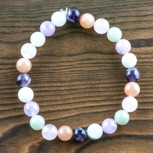 May include: A beaded bracelet featuring a variety of colorful gemstones. The bracelet includes round beads in shades of purple, peach, white, and green. The bracelet is displayed on a dark wooden surface.